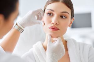 A woman undergoing an aesthetic or dermatologic consultation looks slightly away from the camera as a medical professional examines her face.