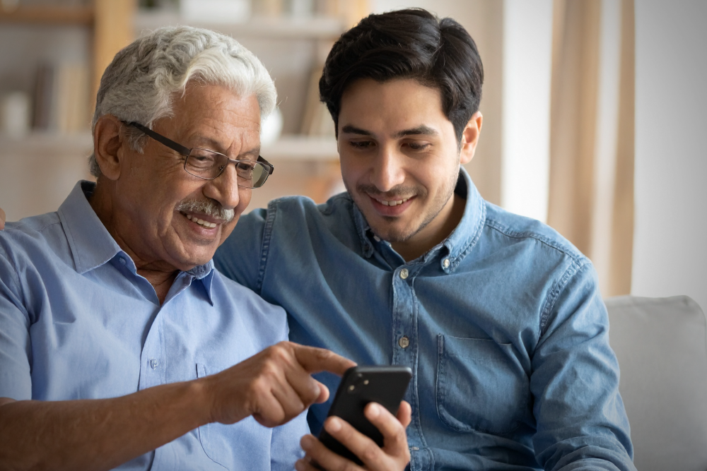 An older man with glasses and gray hair points at a smartphone held by a younger man, both smiling while looking at the screen together in a home setting.